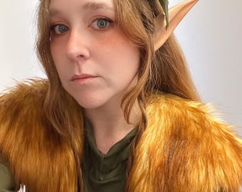 May include: A woman wearing a floral crown with a butterfly accent, faux elf ears, and a faux fur shoulder wrap. The crown features orange, purple, and red flowers. The wrap is a warm, golden brown color. The woman is wearing a green top.
