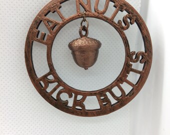 May include: A copper-colored necklace with a circular pendant that reads "Eat Nuts Pick Butts". A small acorn charm hangs from the pendant.
