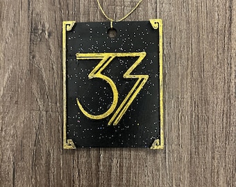 May include: A black rectangular pendant with gold trim and the number "33" in gold. The pendant has a gold string for wearing. The background is a dark wood grain.