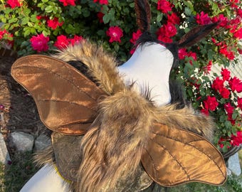 May include: A costume piece featuring brown moth wings with detailed stitching and a faux fur chest piece. The wings are attached to a harness with a decorative, embossed design. The costume is displayed against a backdrop of red roses.