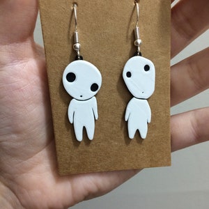 May include: A pair of white dangle earrings featuring a cartoon character with black eyes. The earrings are on a brown card.