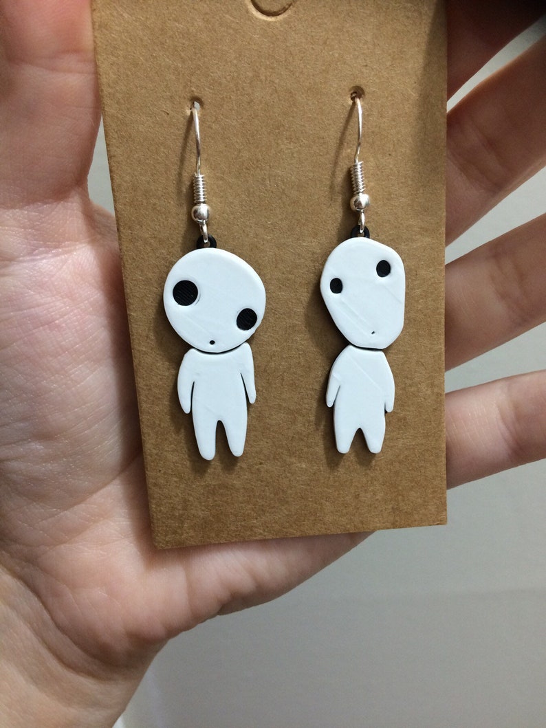 May include: A pair of white dangle earrings featuring a cartoon character with black eyes. The earrings are on a brown card.