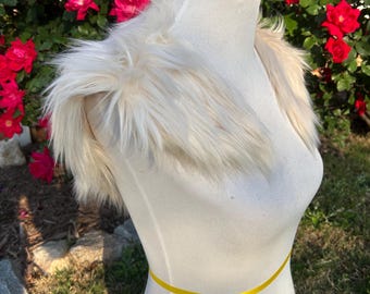 May include: A cream-colored faux fur collar displayed on a white mannequin. The collar has long, soft fur and is designed to be worn around the shoulders. A yellow ribbon is wrapped around the mannequin's waist. Red roses are in the background.