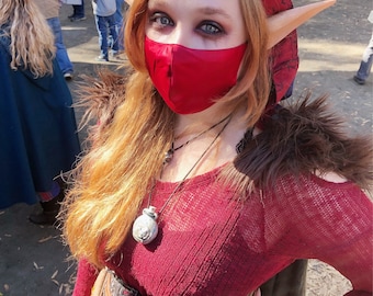 May include: A woman wearing a red hooded sweater with a red face mask, elf ears, and a brown leather belt with a silver buckle. She is wearing a silver necklace with a round pendant.