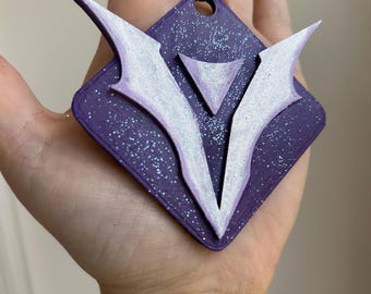 May include: A purple, diamond-shaped pendant with a white and purple emblem. The emblem features a stylized V shape with wing-like extensions. The pendant has a glittery finish and a black string for hanging.
