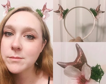 May include: A headband with two antlers and pink flowers. The antlers are made of brown clay and the flowers are made of fabric. The headband is white and has a simple design.