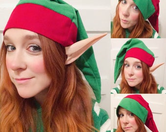 May include: A woman wearing a green and red elf hat with pointed ears. The hat has a red band and a green pointed tip. The woman has long red hair and is wearing a green and white sweater.