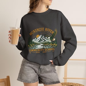 May include: A charcoal gray crewneck sweatshirt featuring a graphic print of a mountain range, a river, a flying eagle, and a sun. The text reads MCKENZIE RIVER COMMUNITY SCHOOL 2026 2027 in gold lettering. The sweatshirt has long sleeves and a ribbed collar.