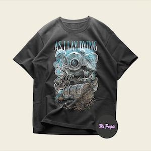 May include: A charcoal gray short-sleeve t-shirt featuring a graphic print of a deep-sea diver in a vintage helmet reaching for a wooden treasure chest. The text AS I LAY DYING is printed in a light blue font above the diver, surrounded by jellyfish.