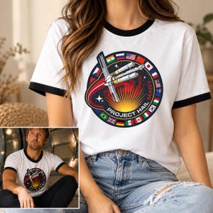 May include: A white ringer t-shirt with black trim on the collar and sleeves. The chest features a circular graphic depicting a spacecraft in space with a sunburst, surrounded by a border of international flags and the text PROJECT HAIL MARY.