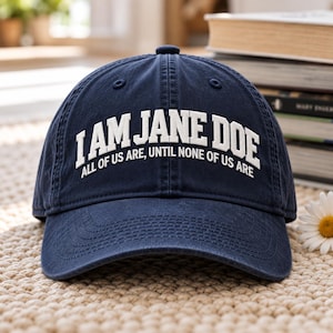 May include: A navy blue baseball cap featuring white embroidered text that reads I AM JANE DOE on the top line and ALL OF US ARE, UNTIL NONE OF US ARE on the bottom line. The cap has a structured crown, a curved brim, and stitched eyelets.