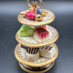 May include: A three-tiered porcelain serving stand featuring gold-toned trim and a central handle. The tiers are decorated with intricate patterns and hold various miniature ceramic food items, including a pink fruit, a green wedge, and several small cakes.