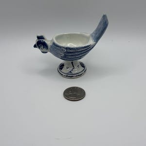 May include: A white ceramic egg cup shaped like a bird with blue painted details on the wings, tail, and base. The bird figure features a small head, a hollow center for an egg, and a pedestal foot. A silver US quarter coin is placed in front.