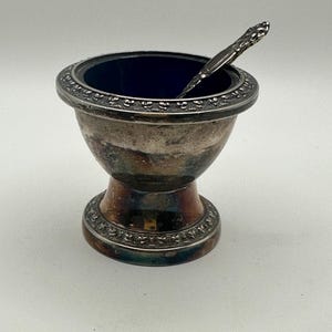 May include: A silver-plated salt cellar with a deep blue glass insert and a matching silver-plated spoon. The bowl and base feature a decorative embossed border pattern. The metal surface shows signs of natural tarnish and oxidation throughout.