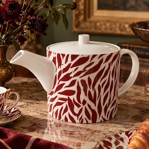 Può includere: Una teiera cilindrica in ceramica con base bianca e un motivo ripetitivo di foglie astratte color rosso scuro. La teiera è dotata di un coperchio bianco con un piccolo pomello, un beccuccio bianco ricurvo e un manico bianco coordinato.