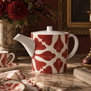 May include: A cylindrical ceramic teapot featuring a white base with a repeating red floral-inspired pattern. The teapot has a white lid, a white handle, and a white spout. A matching teacup and saucer are visible to the left of the teapot.