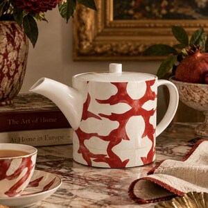 Peut inclure: Une théière cylindrique en céramique blanche ornée d'un motif abstrait rouge répétitif et audacieux. La théière présente une anse incurvée, un bec court et un couvercle plat surmonté d'un petit bouton. Le motif est uniforme sur tout le corps.