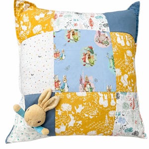 Peut inclure: Un coussin carré en patchwork composé de divers panneaux de tissu illustrés de Pierre Lapin dans des tons de bleu, jaune et blanc. Une figurine de lapin beige en peluche avec des manches bleues est fixée au coin inférieur gauche.