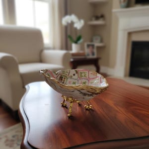 May include: A decorative oyster shell bowl featuring a hand-painted interior with a grid of colorful floral and character motifs. The shell edges are gilded in gold, and the piece is supported by three ornate, polished gold-tone metal legs.