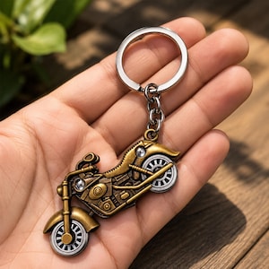 Può includere: Un portachiavi in metallo con un ciondolo a forma di motocicletta dettagliata, caratterizzato da una finitura color bronzo e ruote argentate. La moto è collegata a un anello circolare in metallo argentato tramite una catenella di tre maglie.