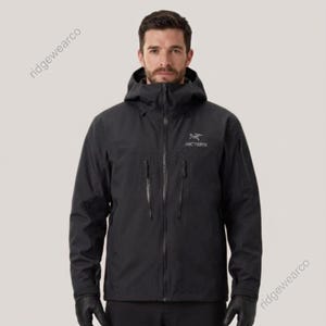May include: A black hooded jacket featuring a full-length front zipper, two vertical zippered chest pockets, and long sleeves. The Arc'teryx logo is embroidered in white on the upper left chest. The jacket has a high collar and a structured hood.