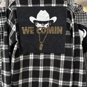 May include: A black and white plaid long-sleeve shirt featuring a large, square, quilted black panel on the back. The panel displays a white graphic of a cowboy hat and sunglasses above the gold-colored text WE COMIN and a gold chain necklace.