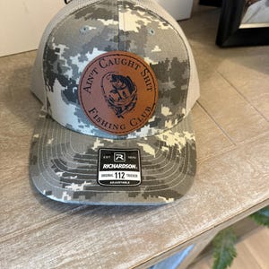 May include: A gray and white digital camouflage trucker hat featuring a circular brown leather patch with a fish graphic and the text AIN'T CAUGHT SHIT FISHING CLUB. A black rectangular sticker on the brim displays RICHARDSON EST. 1970 ORIGINAL 112 TRUCKER.