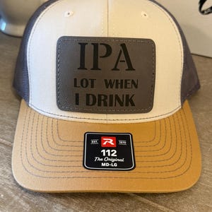 May include: A baseball cap with a cream-colored front panel, dark gray mesh back, and a tan brim. A rectangular dark gray patch on the front features the black text IPA LOT WHEN I DRINK. A black sticker on the brim displays the text R EST 1970 112 The Original MD-LG.