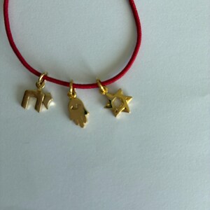 May include: A red cord bracelet featuring three gold-toned charms hanging from the string. The charms include a Hebrew Chai symbol, a Hamsa hand with an eye detail, and a six-pointed Star of David. Each charm is attached with a small gold ring.