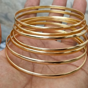 Op de afbeelding: Een set van meerdere dunne, gepolijste armbanden van goudkleurig metaal. Elke armband heeft een glad, afgerond oppervlak met een reflecterende afwerking. De armbanden zijn op elkaar gestapeld en tonen een consistente cirkelvorm en een heldere, metalliek gele kleur.