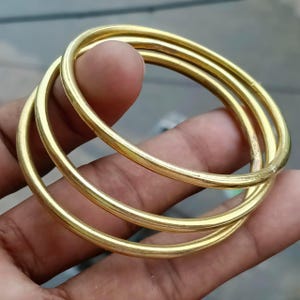 Puede incluir: Un conjunto de tres brazaletes de metal dorado, lisos y pulidos. Cada brazalete presenta un diseño circular simple y continuo con un acabado metálico reflectante. Los tres anillos individuales están apilados formando un conjunto.