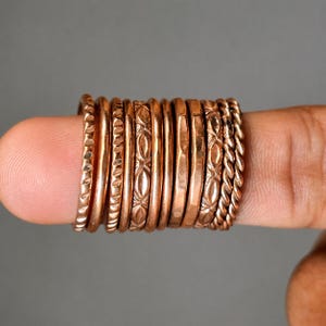 May include: A collection of ten thin copper rings arranged in a row. The rings feature various textures including smooth, hammered, twisted, and patterned surfaces. Each ring has a metallic reddish-brown finish and a circular band design.