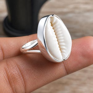May include: A ring featuring a polished silver band and a central setting containing a smooth, off-white cowrie shell. The shell has a characteristic elongated shape with a central slit and ribbed edges, securely mounted within a silver frame.