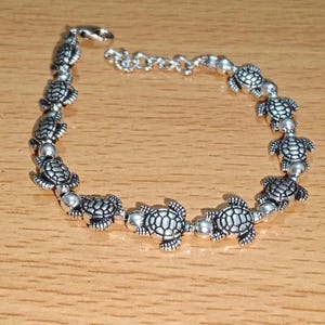 Puede incluir: Una pulsera de metal plateado que presenta una serie de pequeños dijes detallados de tortugas marinas que se alternan con cuentas redondas y lisas de color plateado. La pulsera incluye un cierre de mosquetón y una cadena de extensión.