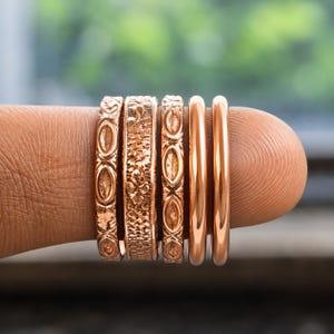 May include: A set of five rose gold-toned rings arranged in a row. The rings feature varied designs, including two plain polished bands, two bands with an embossed oval pattern, and one central band with a detailed, textured floral-like surface.