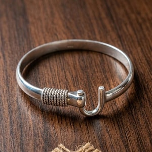 May include: A polished silver bangle bracelet featuring a smooth, rounded band that transitions into a textured, rope-like coiled section near the opening. The bracelet ends in a hook-shaped clasp mechanism, creating a continuous, open-ended circular form.