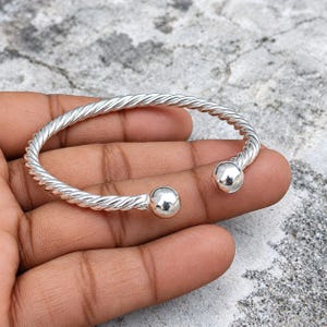 Può includere: Un bracciale a fascia color argento caratterizzato da una texture intrecciata simile a una corda lungo tutto il cerchio. Il bracciale è aperto e termina con una sfera liscia e lucida su ciascuna delle due estremità della struttura metallica.
