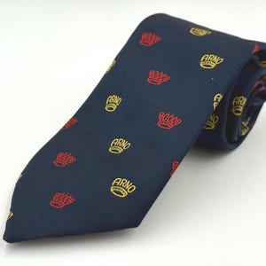 May include: Navy blue tie with a repeating pattern of red crowns and gold "ARNO" text. The tie is folded over itself, showcasing the design. The tie is likely made of a smooth fabric.