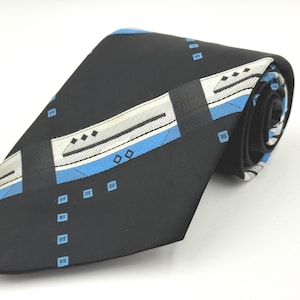 May include: Black tie with a geometric pattern in white, grey, and light blue. The tie features a repeating design of small squares and rectangular shapes. The tie is rolled up, showing the pattern on the back.