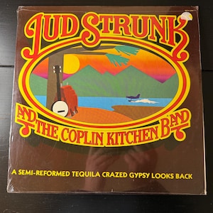 Könnte beinhalten: Ein Vintage-Vinyl-Plattencover mit braunem Hintergrund und einer gelben und orangenen Sonnenuntergangsszene. Der Albumtitel lautet "Jud Strunk and the Coplin Kitchen Band" mit dem Text "A semi-reformed tequila crazed gypsy looks back" unten.