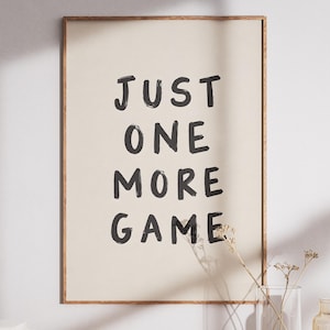 Pode incluir: Uma impressão de arte de parede retangular numa moldura de madeira fina com o texto preto JUST ONE MORE GAME centrado num fundo bege claro. As letras têm um estilo de pincelada pintado à mão com textura e bordas irregulares.