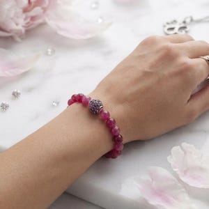 Op de afbeelding: Een kralenarmband met ronde, gefacetteerde, donkerroze edelsteenkralen en een centrale bolvormige bedel ingelegd met kleine, donkerroze steentjes. De armband wordt om de pols gedragen en toont de uniforme grootte van de gepolijste kralen.