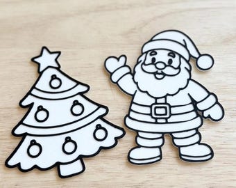Op de afbeelding: Twee platte, wit uitgestanste vormen met dikke zwarte omlijningen. De ene vorm is een kerstboom met een ster op de top en versierd met ronde ornamenten. De andere vorm is een zwaaiende kerstman met een muts, jas en riem.
