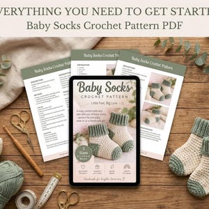 Könnte beinhalten: Ein digitales Tablet mit einer PDF-Häkelanleitung für Babysocken, umgeben von ausgedruckten Anleitungsseiten, zwei Garnknäueln in Creme und Salbeigrün, einer Holzhäkelnadel, einer Schere und einem Paar fertiger Babysocken in Creme und Salbeigrün.