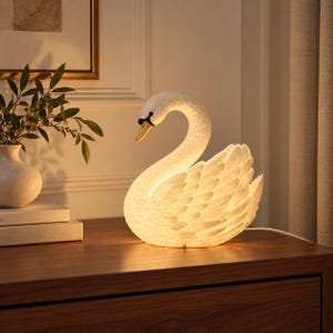 May include: A white swan-shaped table lamp with a textured feather design and a warm internal glow. The swan has a curved neck and a yellow beak with black detailing. The lamp emits a soft light, highlighting the layered plumage of the bird.