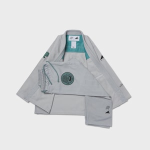 May include: A light gray jiu-jitsu gi featuring a teal inner collar lining and a circular patch on the chest with the letters AP. The garment includes black embroidered logos on the sleeves and pants, with a drawstring waist on the pants.