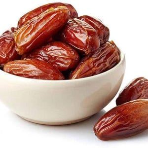 May include: A white ceramic bowl filled with a heap of dark brown, wrinkled dried dates. The dates have a glossy, textured skin and an elongated oval shape. Two additional dried dates are resting on the surface next to the bowl.