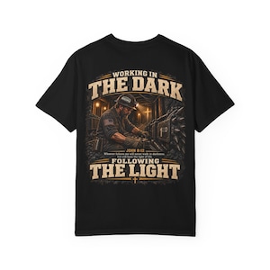 May include: A black t-shirt featuring a graphic print of a miner working in a tunnel. The text reads WORKING IN THE DARK and FOLLOWING THE LIGHT, with the Bible verse JOHN 8:12: Whoever follows me will never walk in darkness, but will have the light of life.