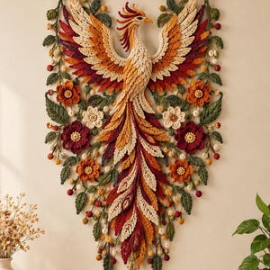 May include: A wall hanging featuring a large crocheted phoenix with wings spread, crafted in shades of cream, orange, and deep red. The bird is surrounded by crocheted green leaves, small yellow berries, and various flowers in red, orange, and cream tones.