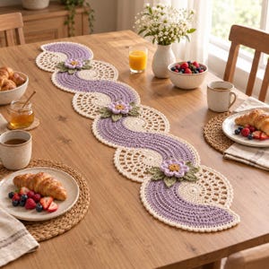 Puede incluir: Un camino de mesa de ganchillo largo y serpenteante con motivos circulares alternos en color lavanda y crema. El camino está adornado con flores de ganchillo tridimensionales de color violeta claro con centros amarillos y hojas verdes.